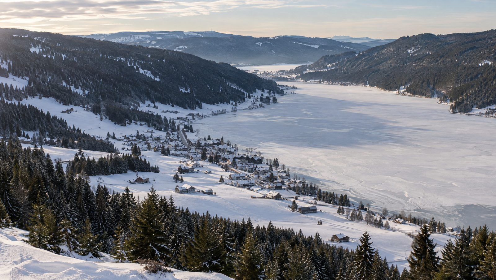 A snow-covered valley in winter, with a frozen lake, a village with a church along its shore and spruce forest on the slopes