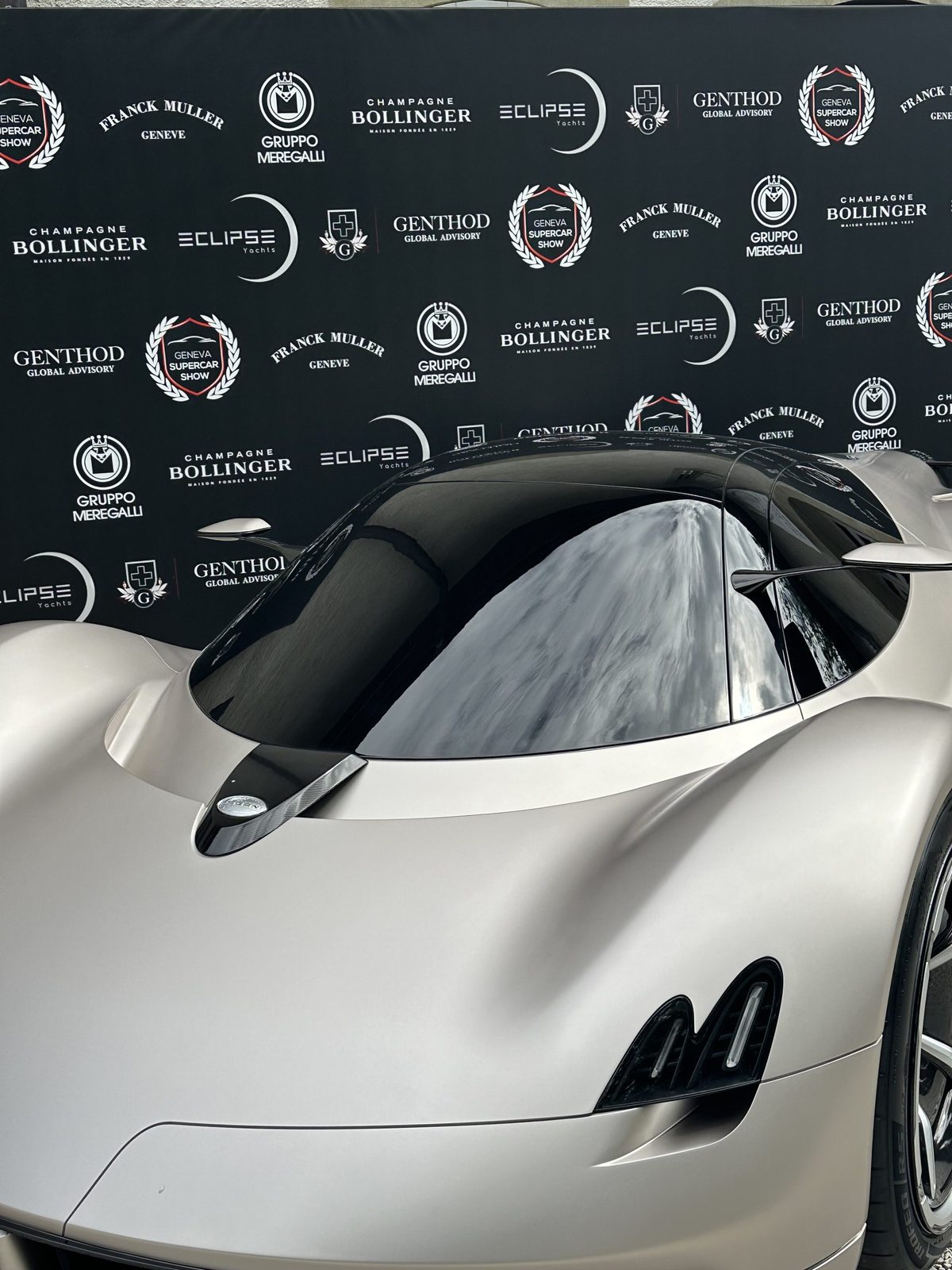 Серебристый прототип гиперкара перед стеной спонсоров Geneva Supercar Show с марками Franck Muller и Bollinger
